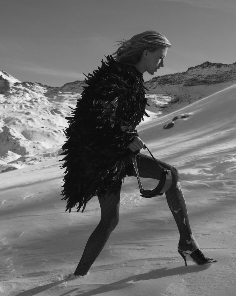 Blonde fashion model wears feather coat and heels while walking on snow in a black-and-white mountain fashion shot.