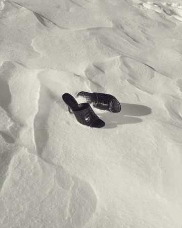 A pair of black croc-embossed heeled mules resting in bright, textured snow with sharp cast shadows.