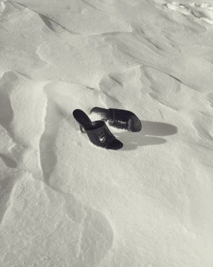 A pair of black croc-embossed heeled mules resting in bright, textured snow with sharp cast shadows.