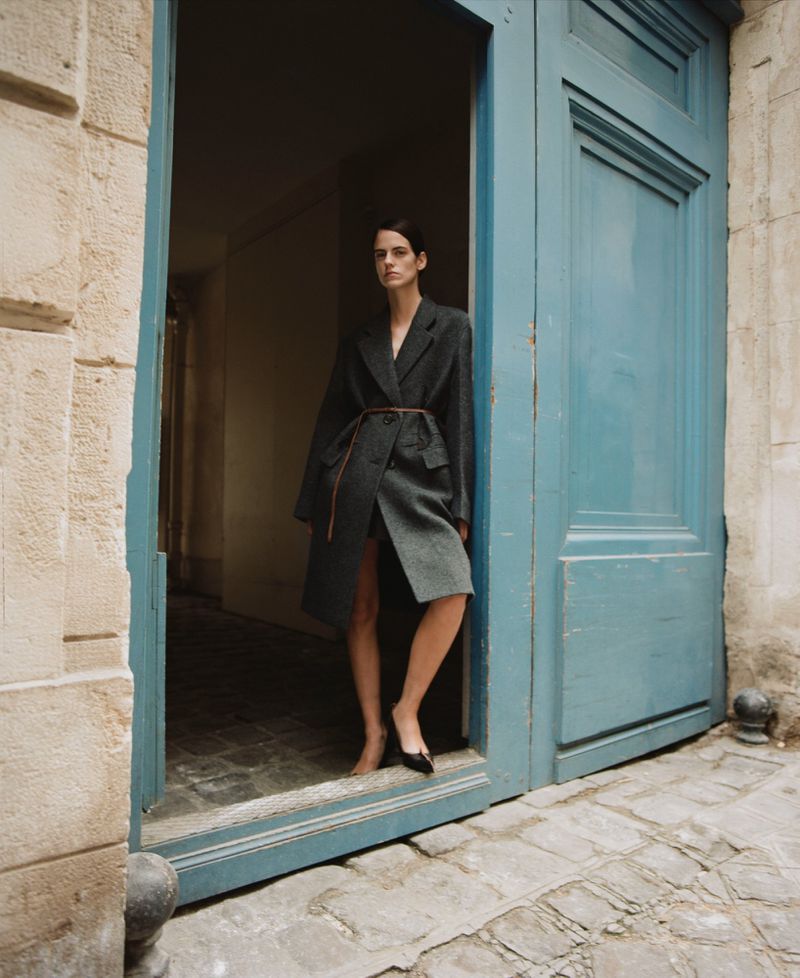A stylish adult woman in a grey overcoat poses confidently in a doorway with blue doors and cobblestone paving in the background.