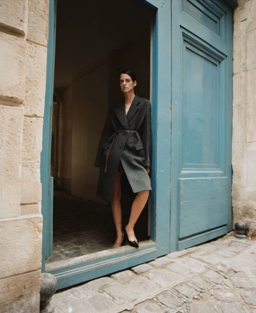 A stylish adult woman in a grey overcoat poses confidently in a doorway with blue doors and cobblestone paving in the background.