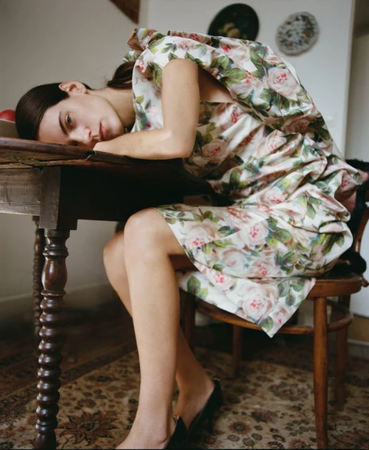 Adult woman in floral dress seated at wooden table with head resting on it, wearing black heels, indoor setting with carpeted floor.