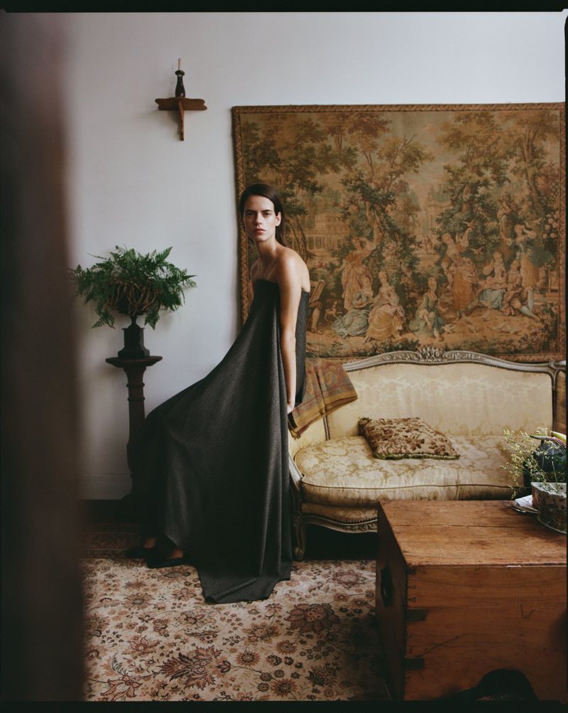 A woman poses indoors wearing a dark, strapless dress that extends to the floor, with a tapestry, sofa, and cabinet in the background.