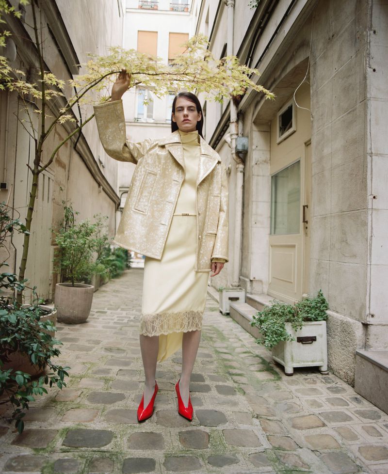 Fashion model poses in a gold jacket, cream dress, and red shoes against a building and foliage-filled alley in an outdoor setting.