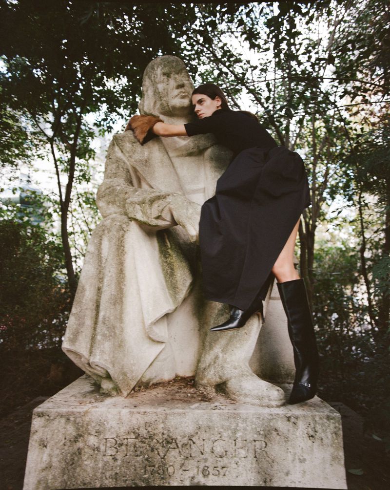 A woman in black attire embraces a stone statue in an outdoor park setting, amidst trees and foliage, under natural lighting.