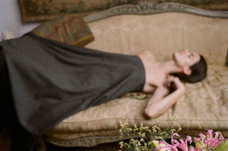 Adult female lies on a beige floral couch wearing a black dress with flowers in the foreground in an indoor setting.