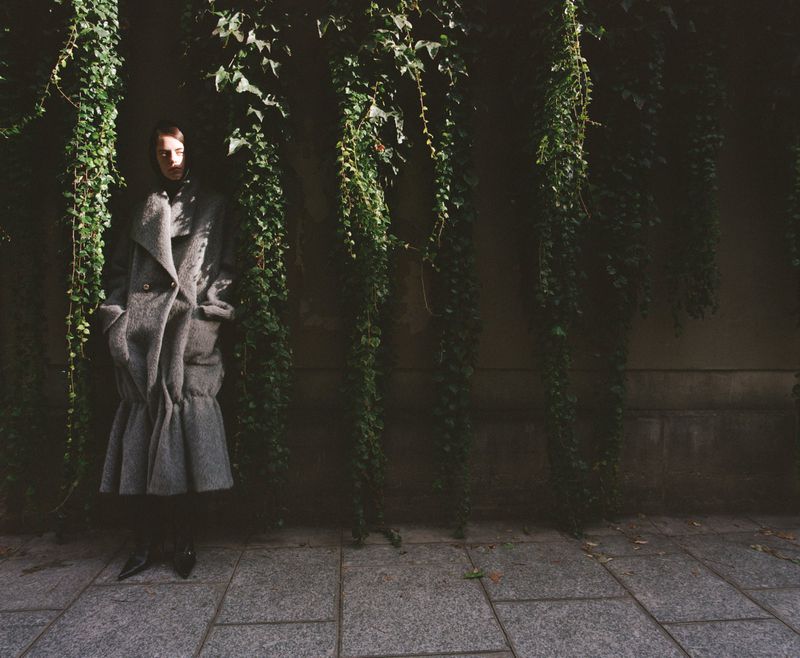Woman in a grey wool coat standing on a stone path in front of a vine-covered wall, captured in a full shot on an overcast day.
