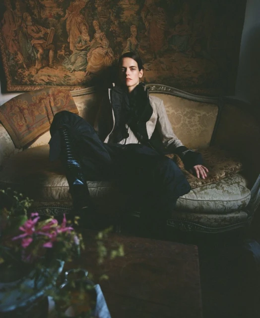 Woman in jacket and dark pants lounges on antique sofa beneath tapestry in indoor setting with flowers in foreground.