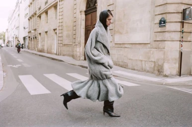 Model Miriam Sanchez running across a city crosswalk wearing an oversized grey flared coat, black headscarf, and black heeled boots.