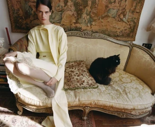 Model in pale yellow dress seated on an ornate sofa with a black cat for Twin Magazine