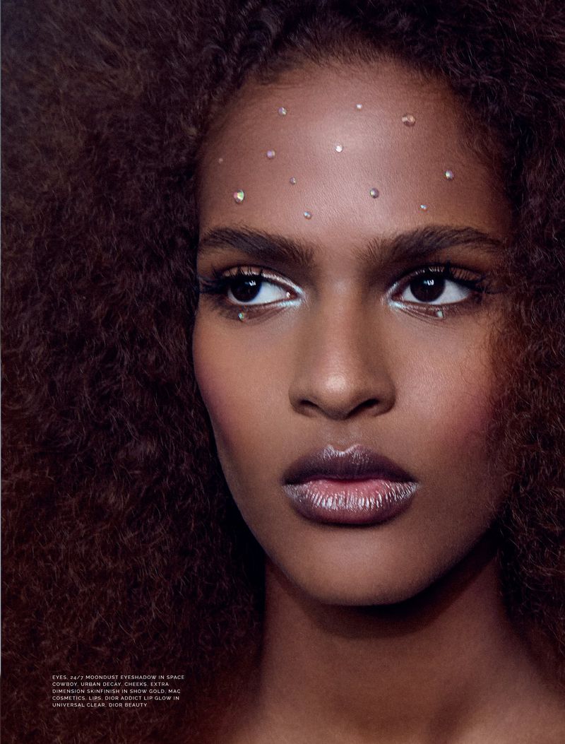 Close-up portrait of a Black woman with an afro, wearing dramatic white eye makeup and iridescent lipstick, with gems on her forehead.
