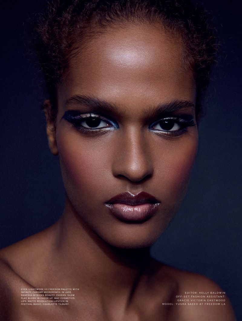 Close-up portrait of a woman with dramatic blue eye makeup and glossy lips against a dark background in a beauty fashion editorial.