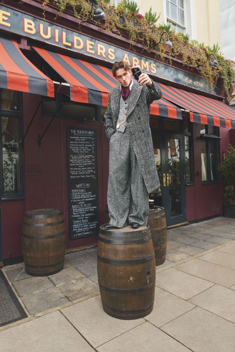 A man in a grey coat standing on a wooden barrel outside of The Builder's Arms pub, holding a pint of beer toward the camera.