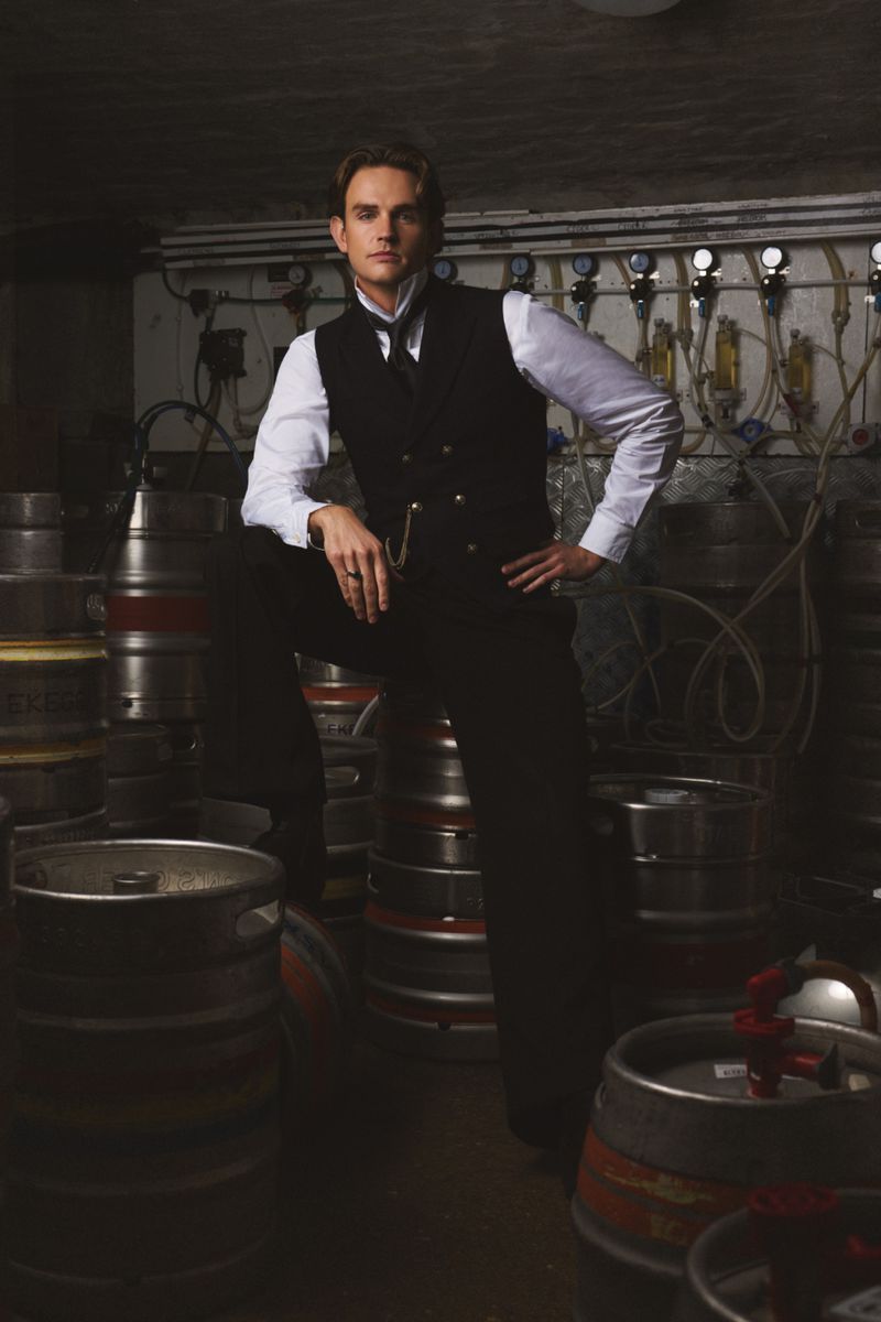 Man in a vintage vest poses in a cellar-like room with stacks of metal kegs and industrial pipes, creating a classic aesthetic.