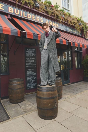 Man in an oversized grey tweed suit and overcoat standing atop a wooden barrel outside a pub while taking a selfie on his smartphone.