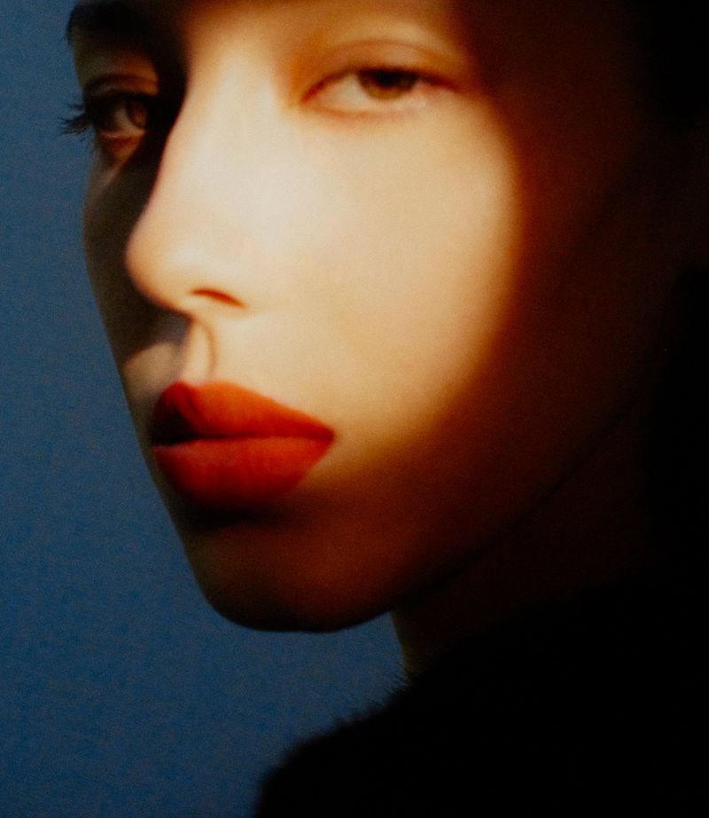 Close-up portrait of a young adult woman with red lips and fair skin, set against a dark blue background with a golden light.