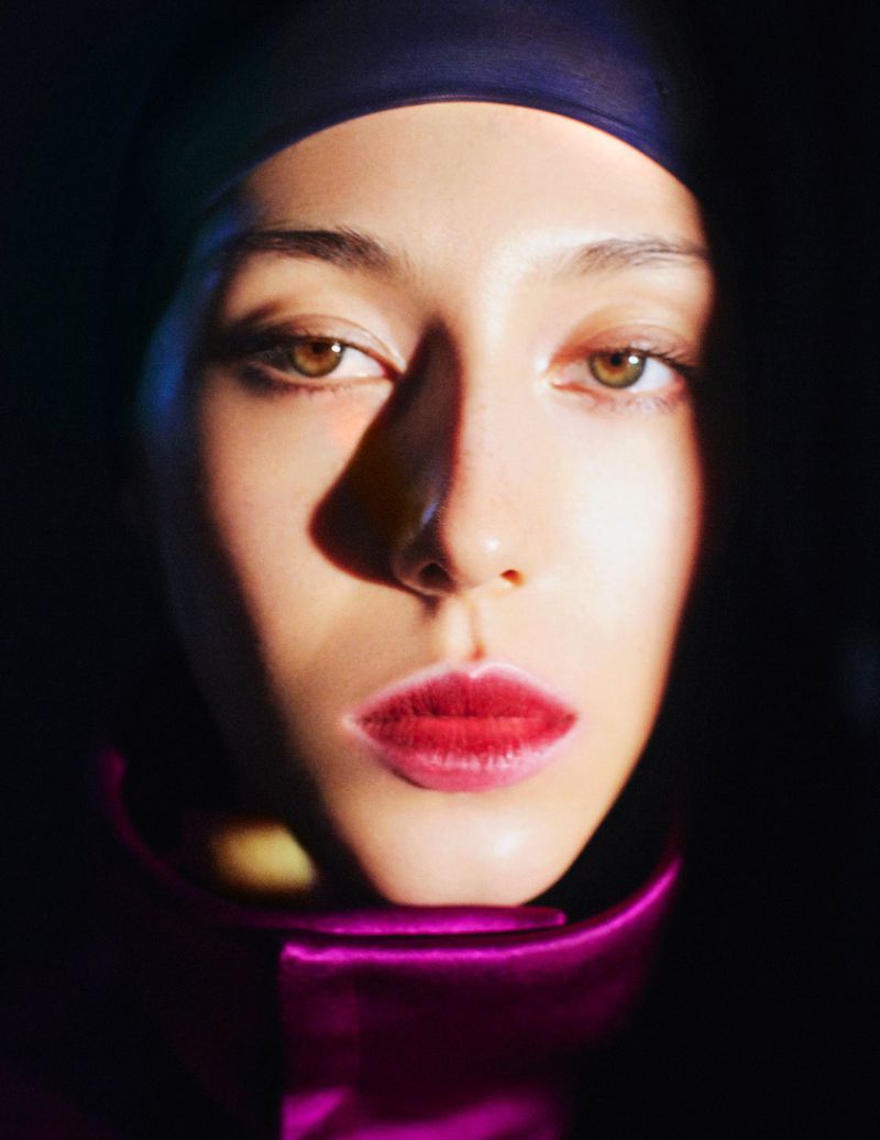 Close-up portrait of a woman with red lipstick and a blue head covering, set against a dark background with a ray of light on her face.