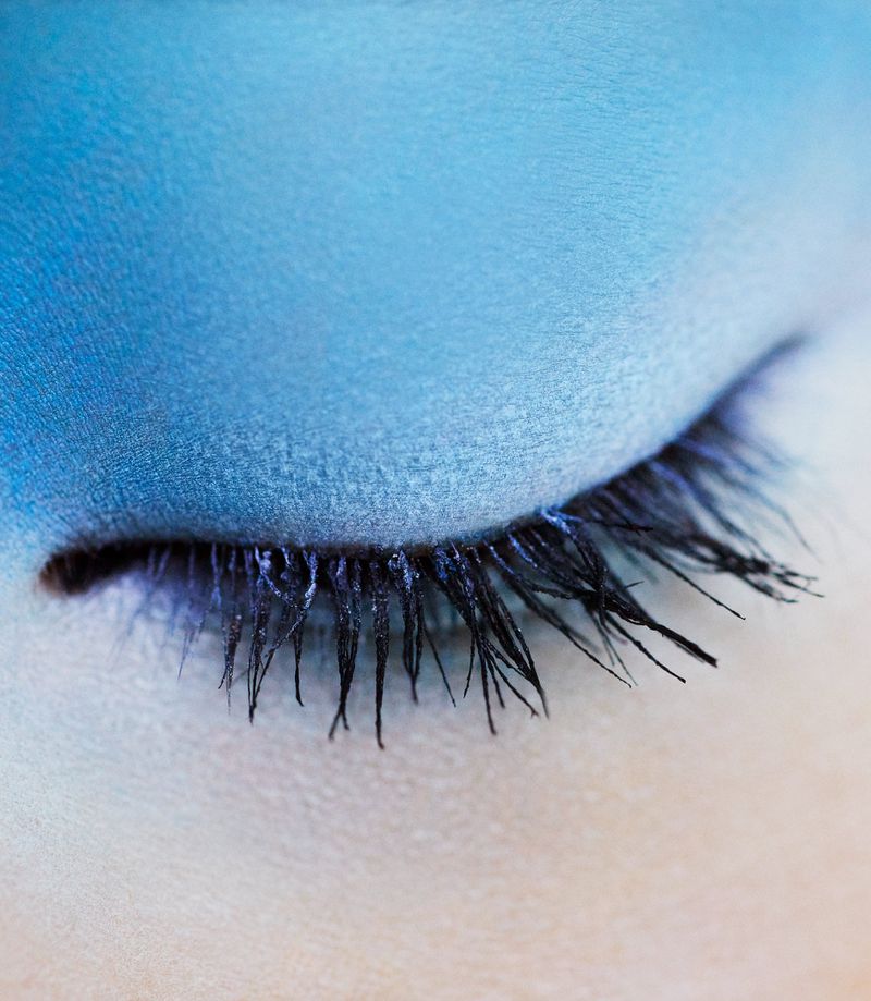 Macro photograph of a closed eye with dramatic blue eyeshadow and long, dark eyelashes, showcasing detailed makeup artistry.