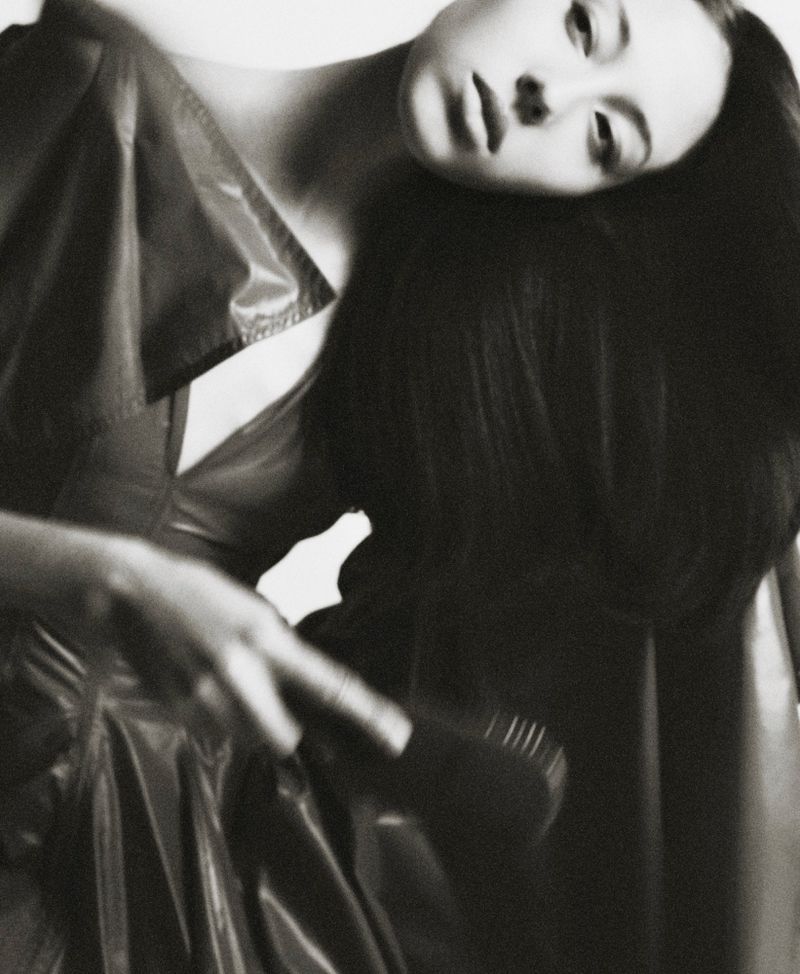 Woman with long dark hair brushes her hair indoors with a blurred-motion brush, styled in a black-and-white editorial fashion photograph.