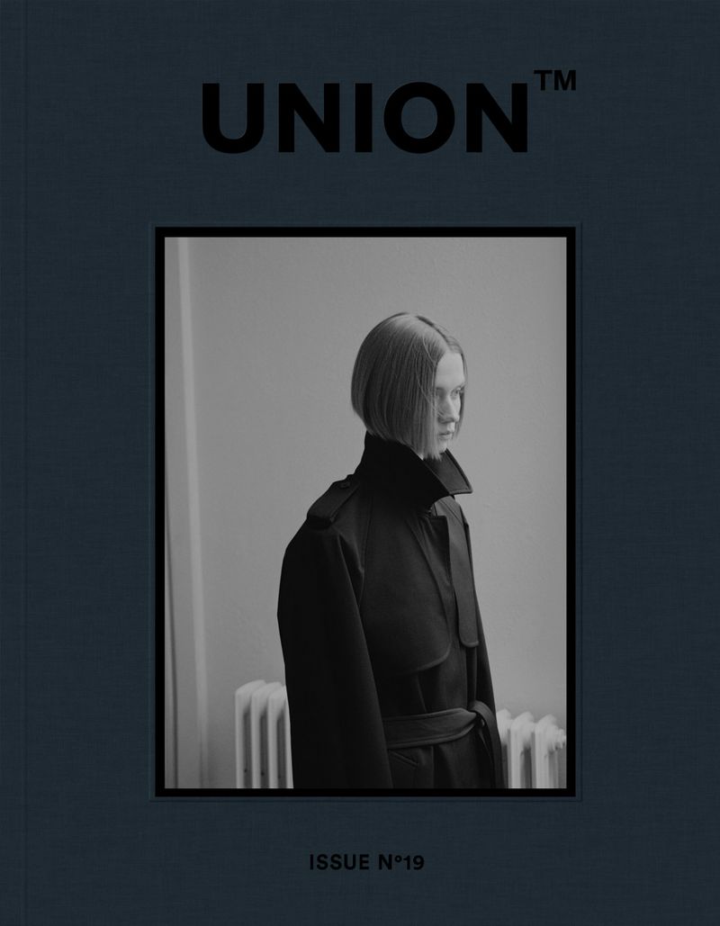 Black and white portrait of a model on a magazine cover. The model has short blonde hair and is wearing a black coat with a belt.