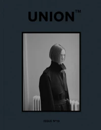 Model in a black trench coat posing against a plain wall for Union Magazine N° 19 editorial photography