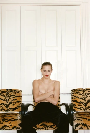 A striking portrait of a topless woman with dark pants, seated between tiger-print chairs against a white paneled wall, fashion style.