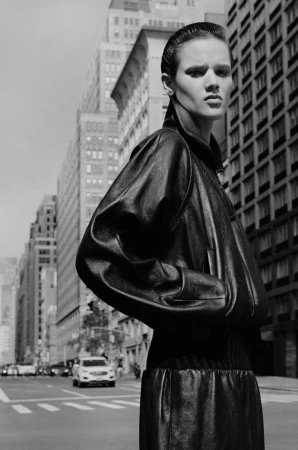 Monochrome portrait of a fashion model in a black leather jacket and skirt, standing on a city street with buildings in the background.
