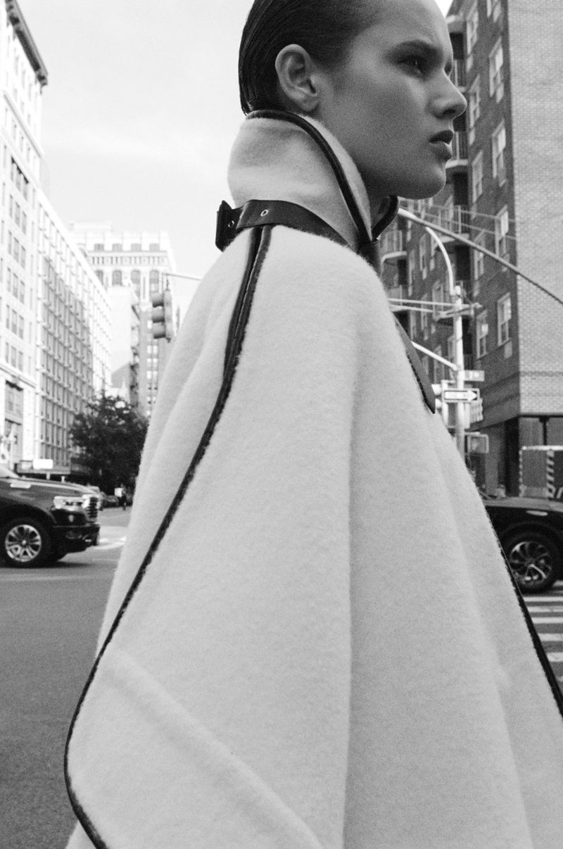 Black and white portrait of a woman wearing a light-colored poncho with dark trim on a city street with buildings and cars.