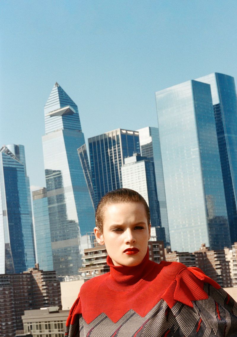 Fashion shot of a woman with short hair wearing a red and grey top against a backdrop of urban skyscrapers under a bright sky.