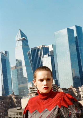 Fashion shot of a woman with short hair wearing a red and grey top against a backdrop of urban skyscrapers under a bright sky.