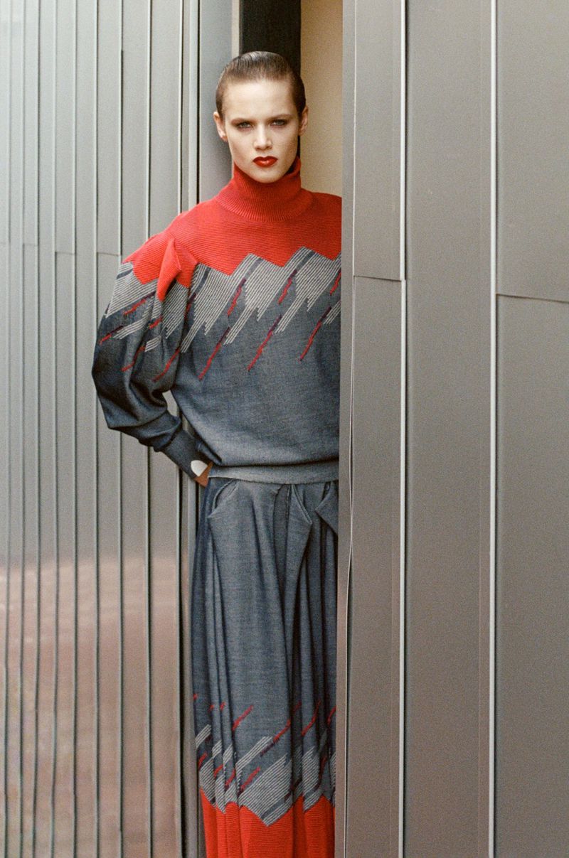 Adult female fashion model posing in a red and gray dress outside, standing next to a wall of metal panels in natural light.