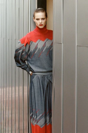 Adult female fashion model posing in a red and gray dress outside, standing next to a wall of metal panels in natural light.