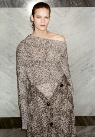 A slender adult female poses in a one-shoulder, button-up dress made of textured gray and brown fabric against a marble wall.