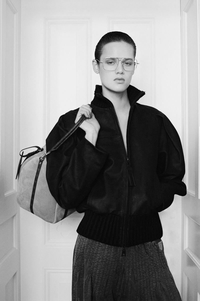 Monochrome portrait of a woman wearing a black bomber jacket and glasses, carrying a light-colored duffel bag in an indoor setting.