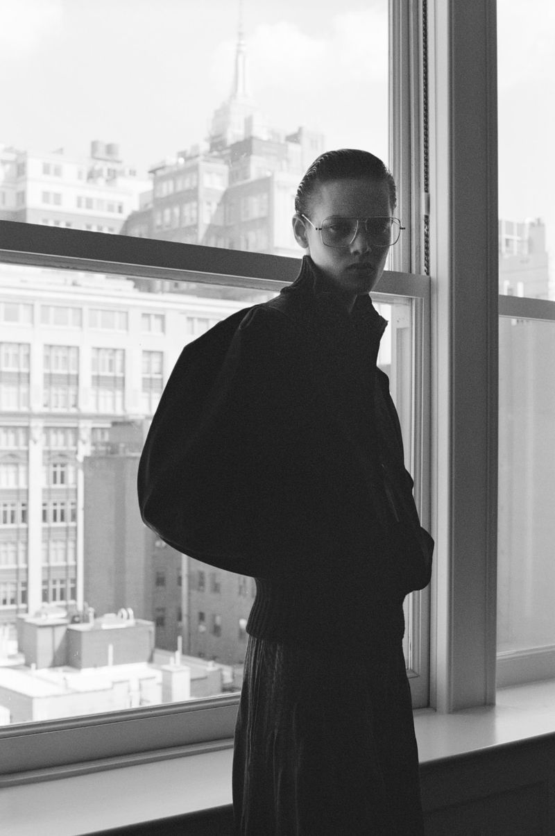 Monochrome portrait of an adult posing near a window, with city buildings visible in the background, creating a stylish and urban scene.