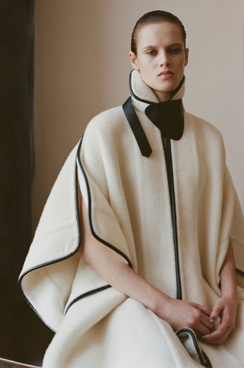 Indoor shot of a poised, adult woman wearing a white cape with black trim, accessorized with a black high-neck collar, against a plain backdrop.