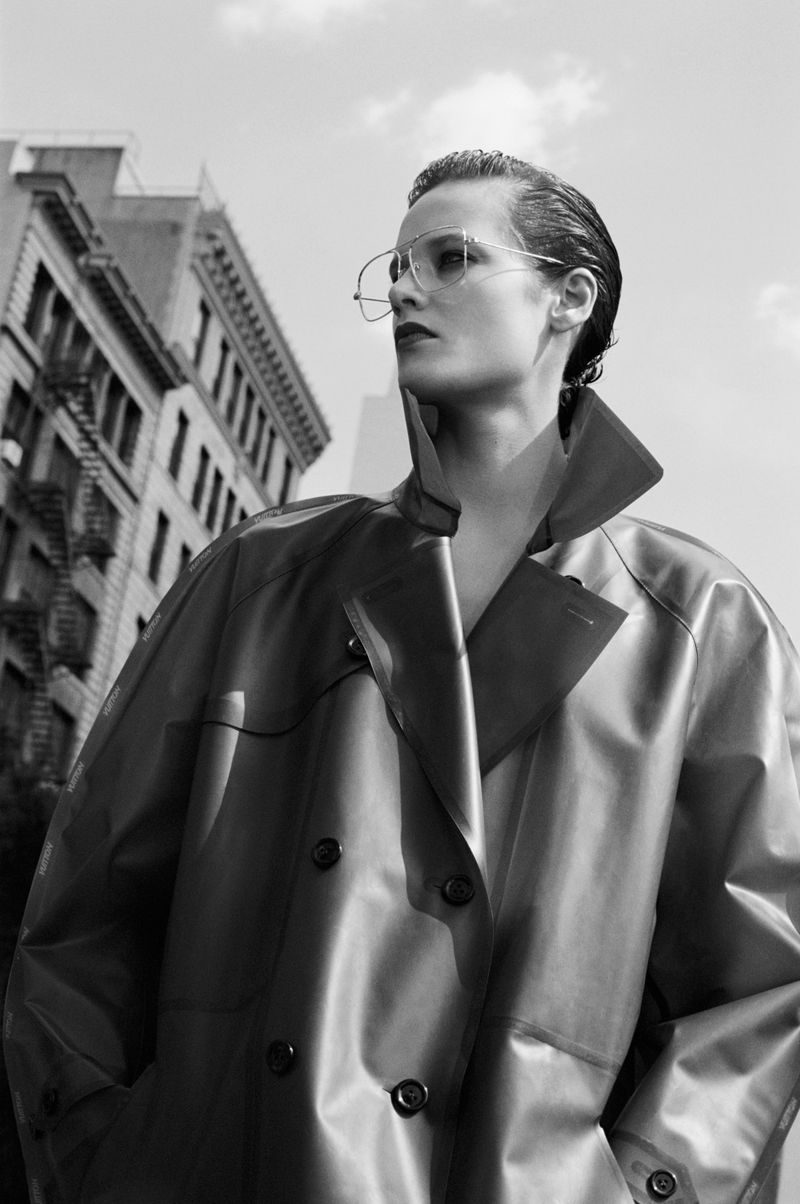 A stylish adult in an oversized trench coat and aviator glasses stands against a backdrop of city buildings under a cloudy sky.