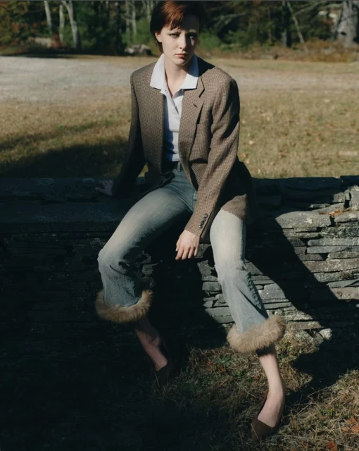 A woman with red hair sits on a stone wall wearing a blazer, white collared shirt, cuffed denim jeans, and brown shoes in an outdoor setting.