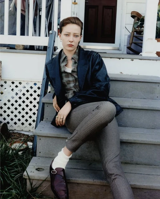 Woman sits on porch steps wearing a jacket, plaid shirt, pants, socks, and a loafer in an outdoor setting with greenery behind.