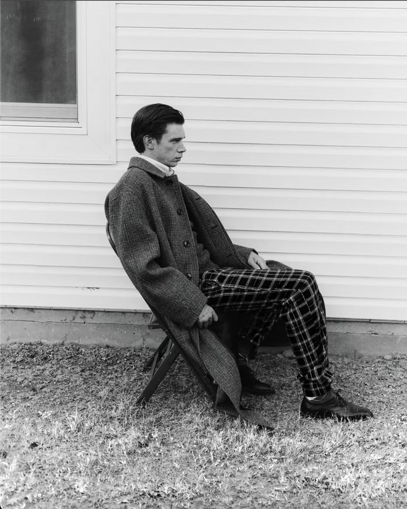 Monochrome portrait of a young adult male seated outdoors, wearing a coat and patterned trousers, against a light-colored wall with a window.