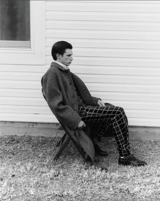 Monochrome portrait of a young adult male seated outdoors, wearing a coat and patterned trousers, against a light-colored wall with a window.