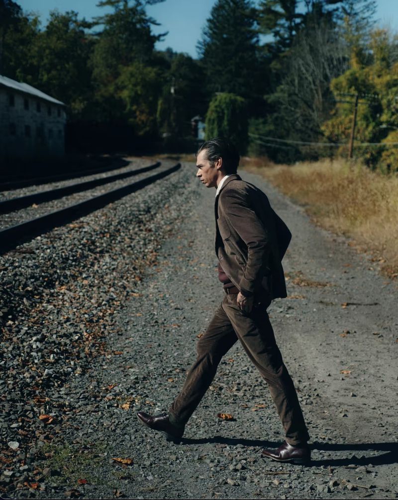 A man in a brown suit walks across a gravel path with train tracks, backed by a building and lush trees under a blue sky.