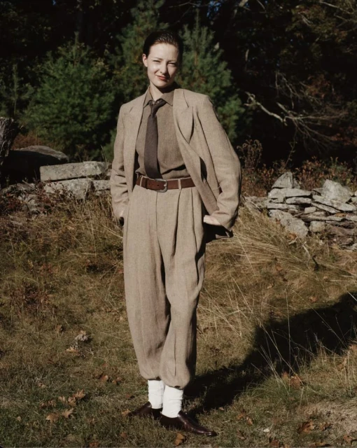 A woman stands outdoors in tailored brown suit with a tie, complemented by brown leather shoes and white socks against a natural backdrop.