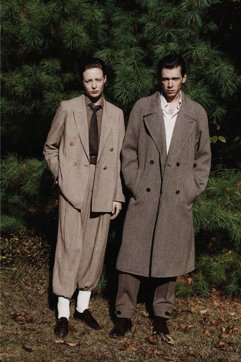 Two people stand outdoors wearing neutral-toned vintage-style coats and suits against a backdrop of green foliage and dappled light.
