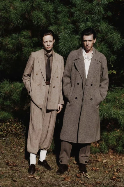 Two people stand outdoors wearing neutral-toned vintage-style coats and suits against a backdrop of green foliage and dappled light.
