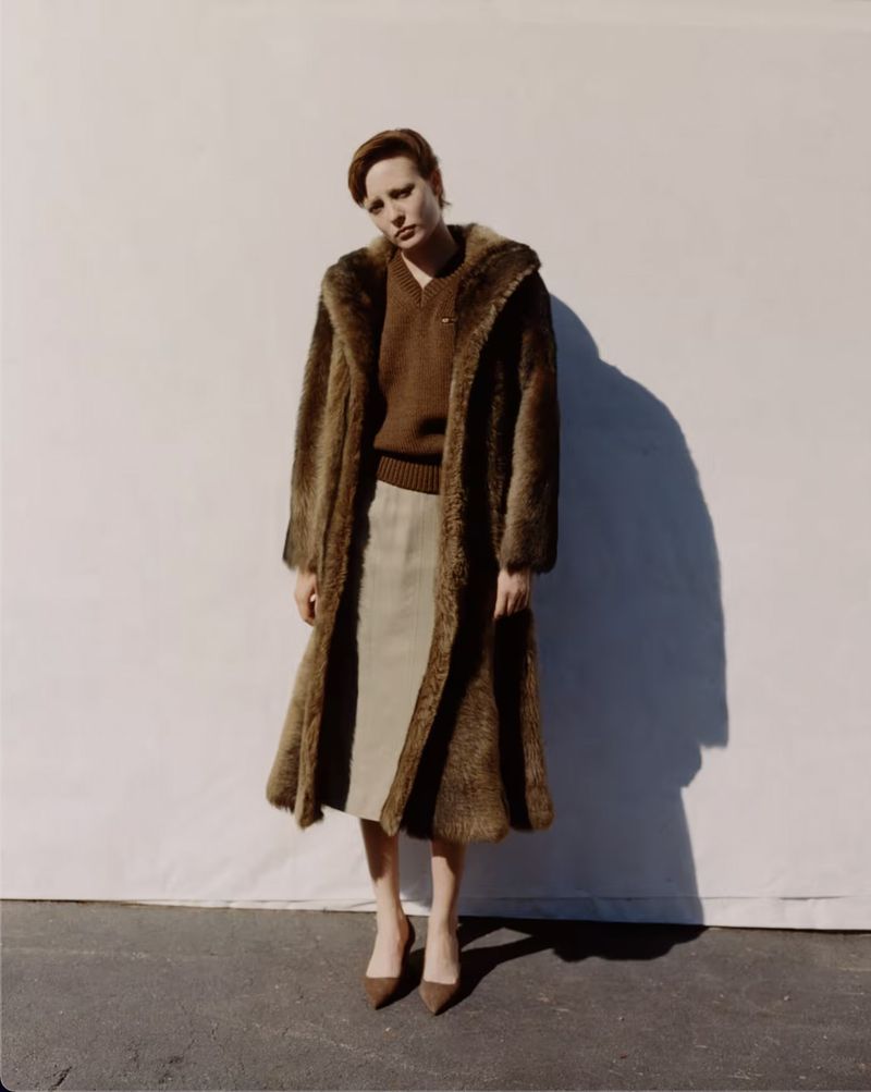Model wearing a brown fur coat over a knit sweater and skirt against a pale backdrop
