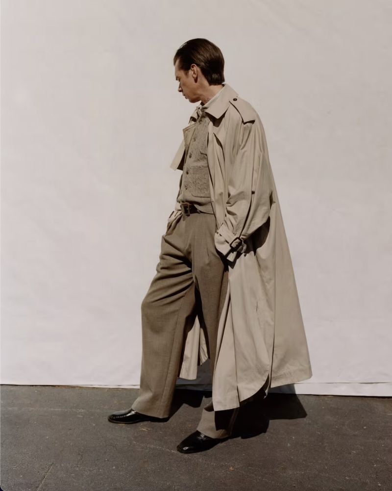 A man wearing a tan trench coat, knit vest, and trousers posing against a neutral studio backdrop.