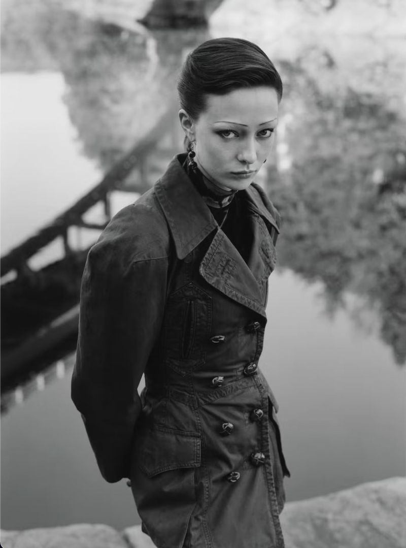 Black and white portrait of a young woman in a trench coat, posing outdoors near a river with a bridge reflection in the background.