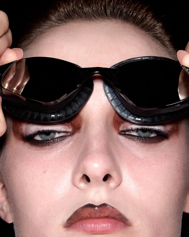Close-up studio shot of a model with dark eye makeup adjusting oversized black goggles on her face against a black background.