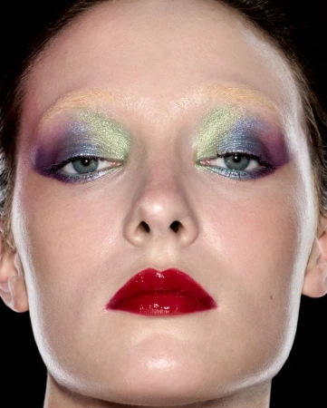 Close-up studio portrait of a person with bold, colorful eye makeup and glossy red lips against a black background.